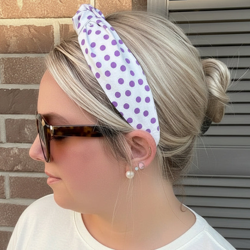 A woman wearing a white fabric headband with purple polka dots, styled in a side profile against a brick wall and shuttered window.