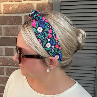 A woman wearing a wide fabric headband with a colorful floral print, styled in a side bun. The headband features a navy blue background with pink, green, and white flowers.