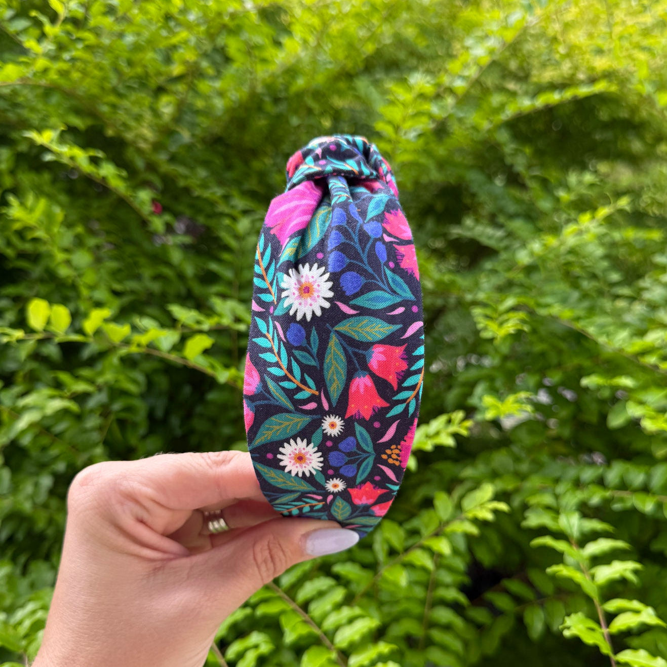 A hand holding a small fabric drawstring bag with a colorful floral print against a background of green bushes.