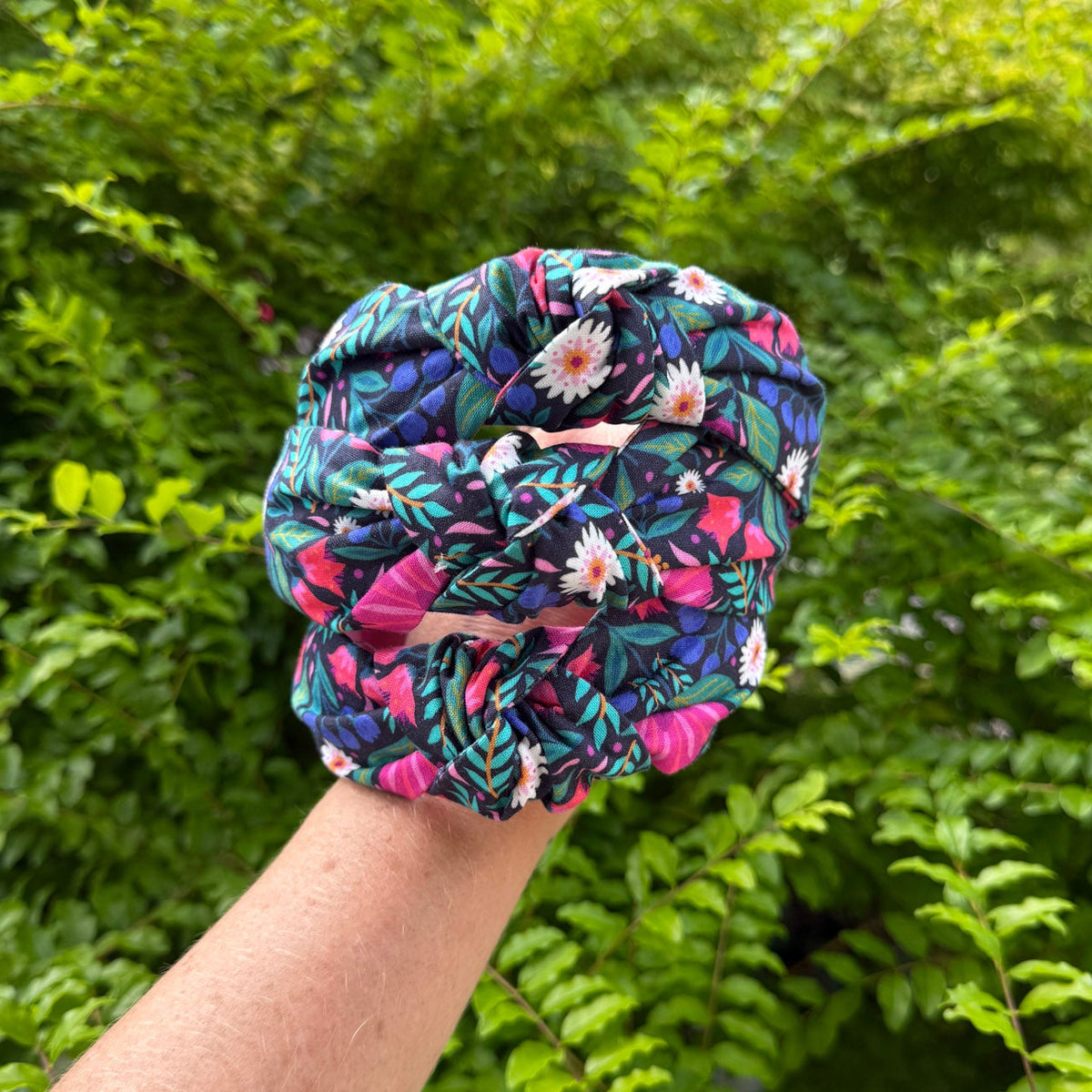 A fabric hair scrunchie with a colorful tropical floral and leaf print, held up by a person's hand against a background of green bushes.