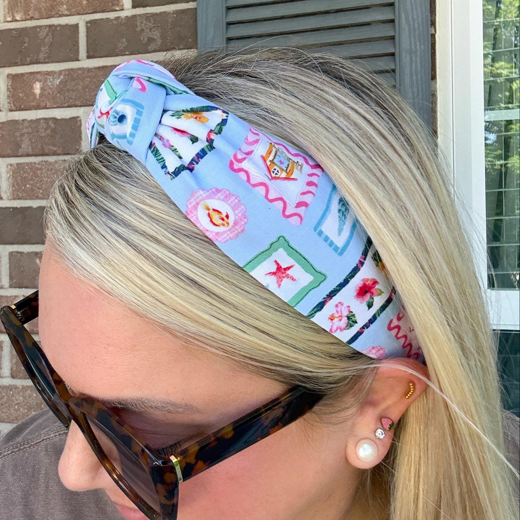 A blonde woman wearing a light blue patterned fabric headband, tortoiseshell sunglasses, and a brown shirt, shown in profile against a brick wall and window background.